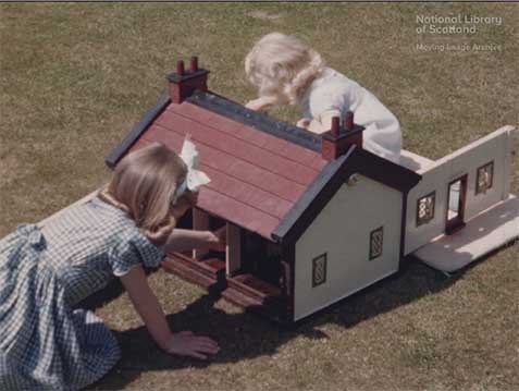 Two young girls playing with a doll's house in a garden on a sunny day.