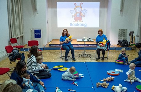 Two people sitting on chairs holding bookbug soft toys. Other people with babies are sitting on blue mats on the floor.