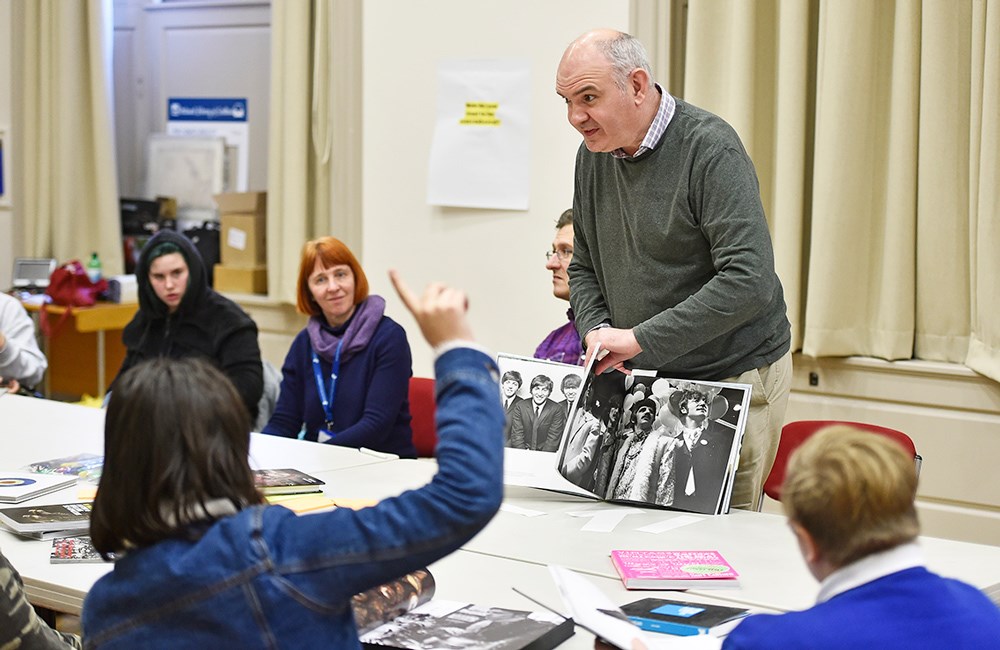 A member of staff standing and showing visitors a book with black and white photos of the Beatles.