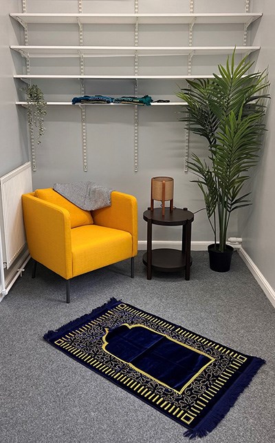 A photo of a room. There is an armchair, with a small table, lamp, plant, and prayer rug on the floor.