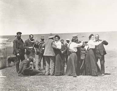 A black and white photo of men and women in old dress dancing and playing instruments outside.