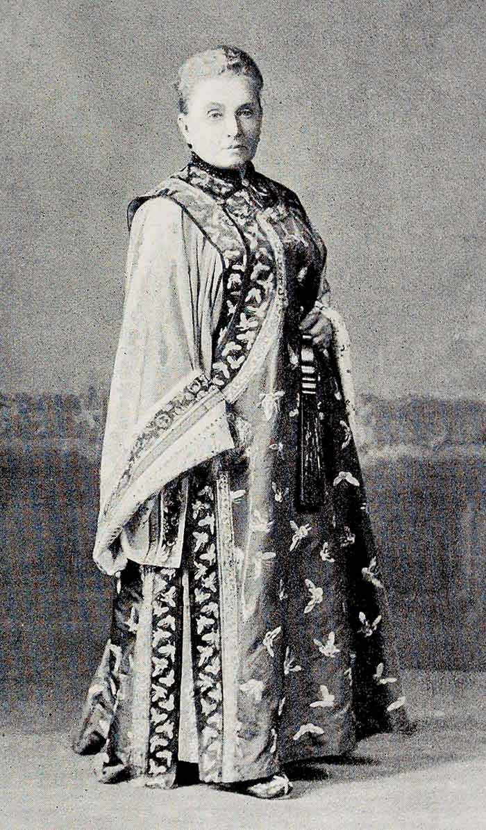 A black and white photograph of Isabella Bird dressed in tradition clothing standing up.