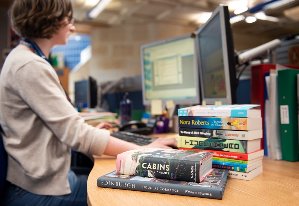 Person working at a desk with a pile of books in the foreground.