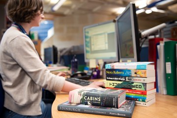 Person working at a desk with a pile of books in the foreground.