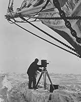 Frank Hurley on the ice with a camera, alongside the ship