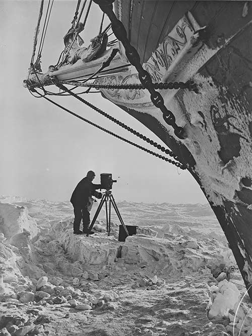 Frank Hurley on the ice with a camera, alongside the ship