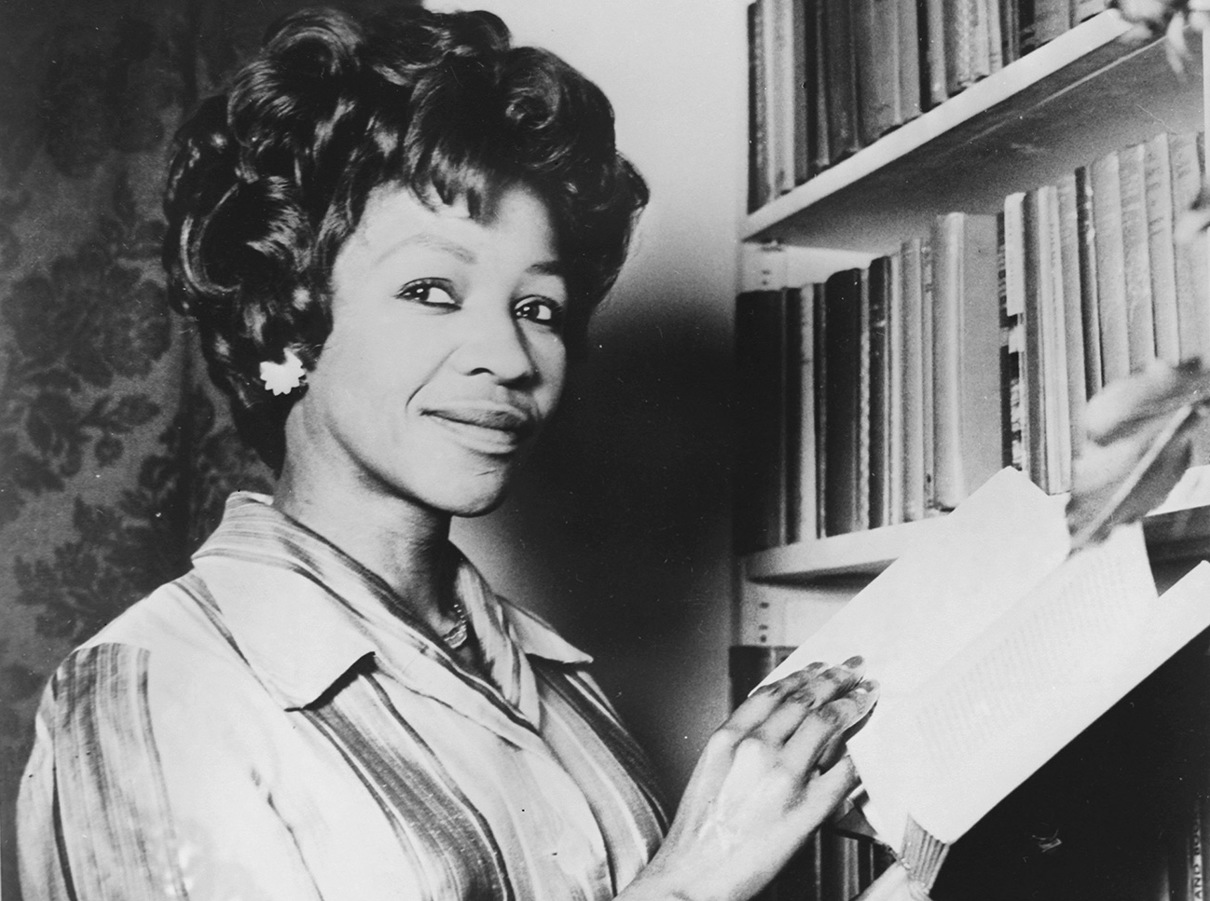 Black and white photo of Noni Jabavu standing in front of a bookcase holding a book.