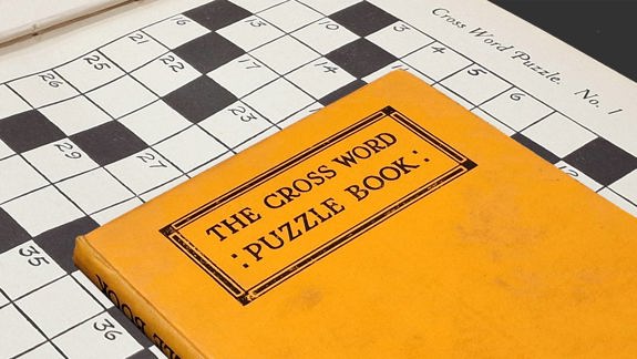 An orange crossword puzzle book superimposed on one of the black and white crosswords within.