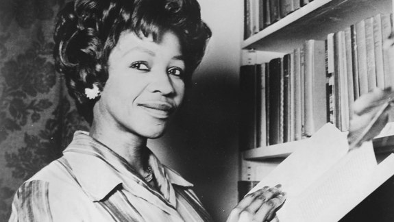 Noni Jabavu standing in front of a bookcase holding a book.