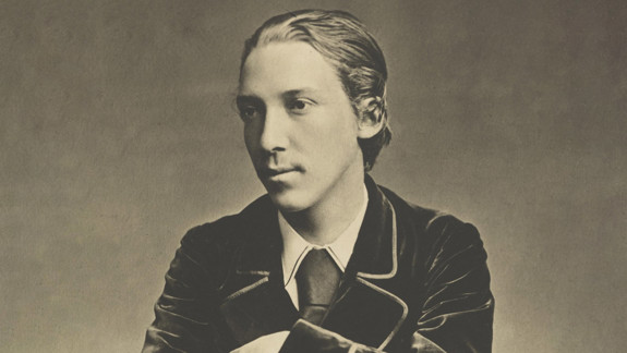 Sepia-toned photograph of a man sitting on a chair with arms crossed over the backrest. The person wears a dark velvet jacket, white shirt, and tie.