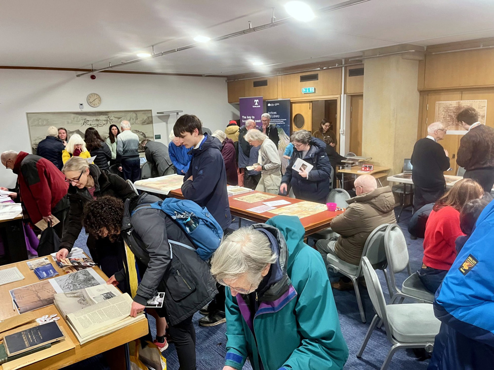 Lots of people in a room looking at maps and other archival material on display on tables.