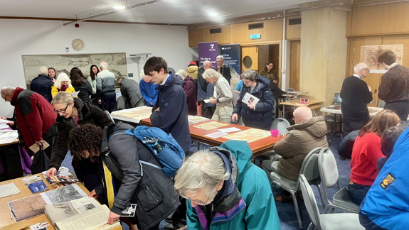 Lots of people in a room looking at maps and other archival material on display on tables.