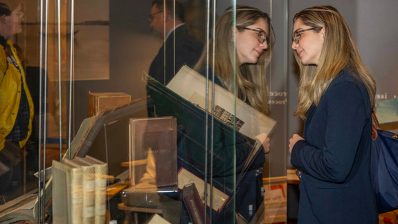 A person standing looking at an exhibition. Their image is reflected in the exhibition case.