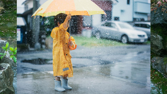 A young boy standing in the rain wearing a yellow raincoat and holding an umbrella.