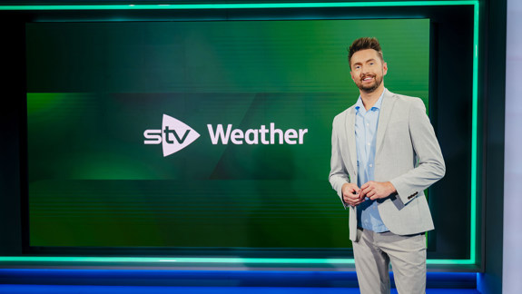Person standing in a television studio beside a screen displaying the STV Weather logo.
