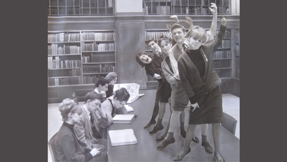 A black and white photo of people sitting at a desk reading and then standing on the desk posing.