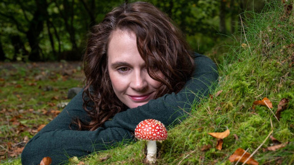 A woman lying on a woodland floor looking at a red and white mushroom.