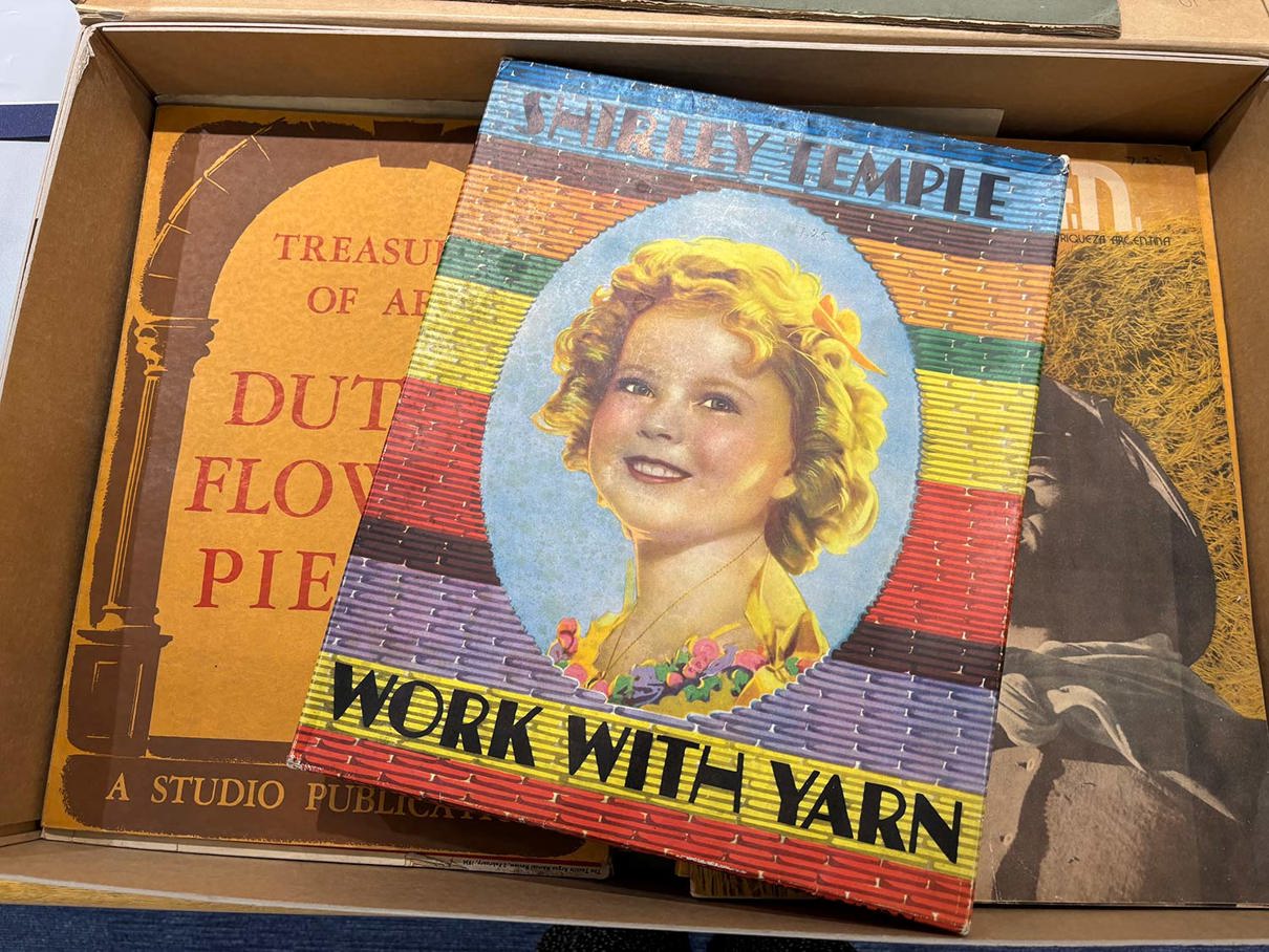 A box containing three vintage books and other items. The top item is a box titled Shirley Temple Work with Yarn, featuring an illustration of a blonde girl.