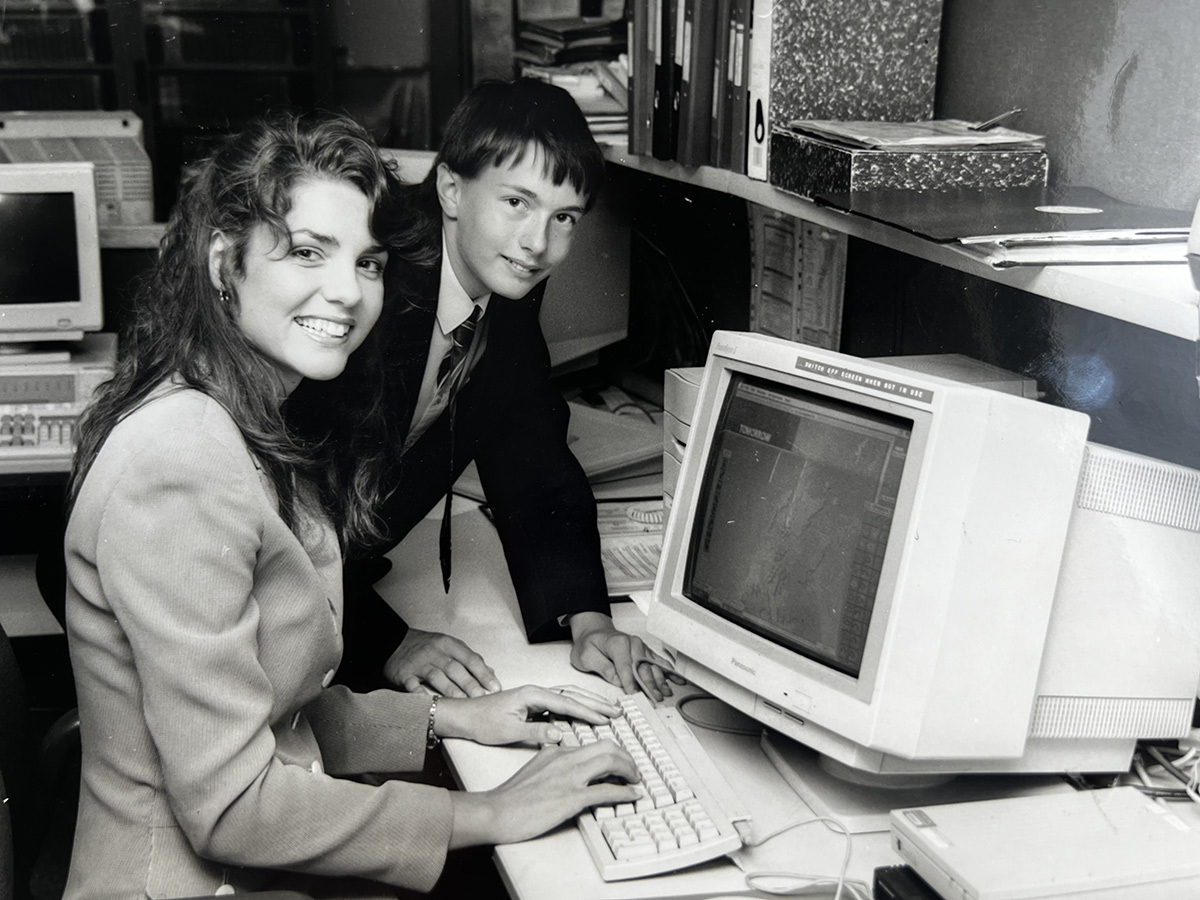 Black-and-white photograph of two people using a desktop computer in an office, with shelves and files in the background.