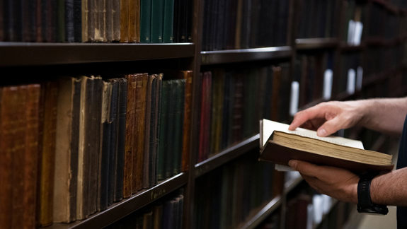 A person with an open book reading in front of large library stacks with lots of old books