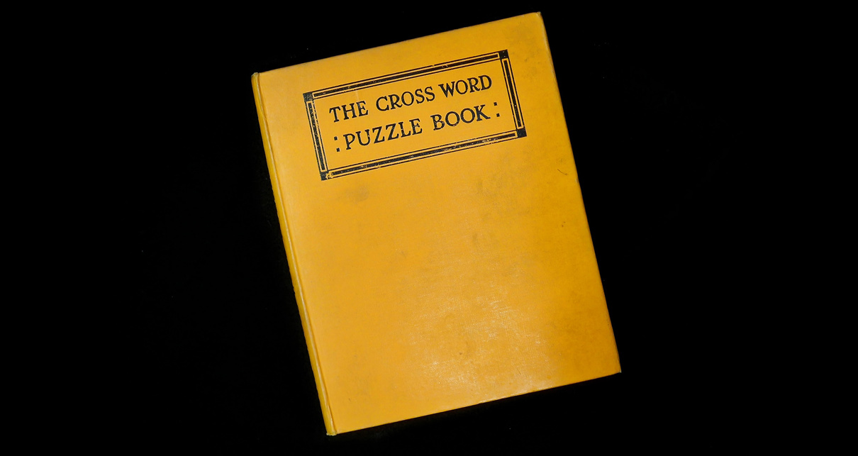 An orange book against a black background. The book cover has "The Cross Word Puzzle Book" printed on it in black.