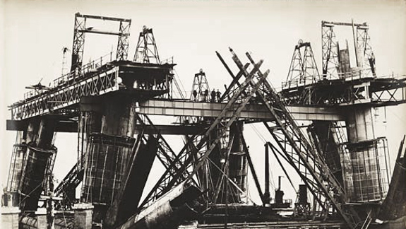 Black and white photo of a large construction site with steel beams, cranes, scaffolding, and workers building a bridge.