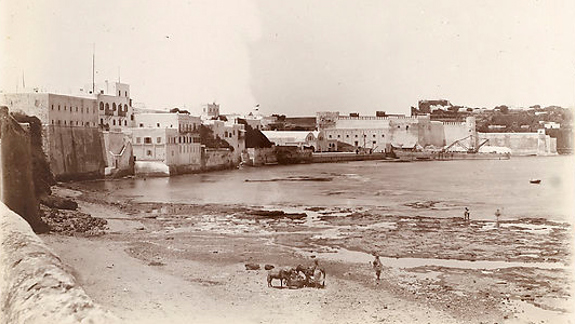 A coastal scene featuring historical multi-story buildings with arches and balconies along the shoreline. A sandy beach with a few people is in the foreground, and calm water with rocky areas is visible.