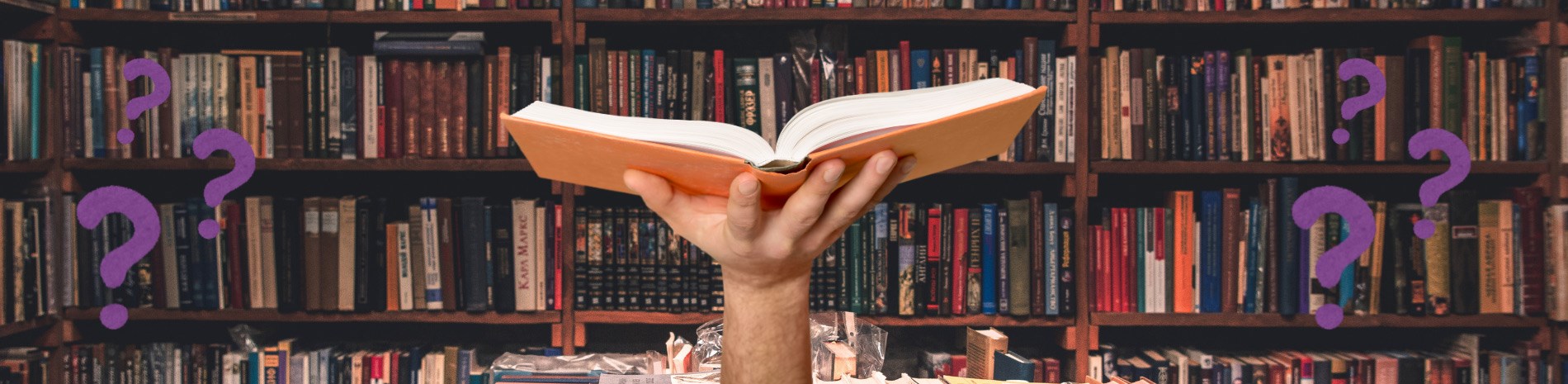 A hand holding up an open book with shelves full of books in the background and question marks around.