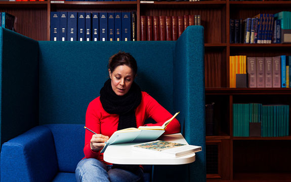 A reader is seated in a blue armchair and writing in a notebook while looking at a book