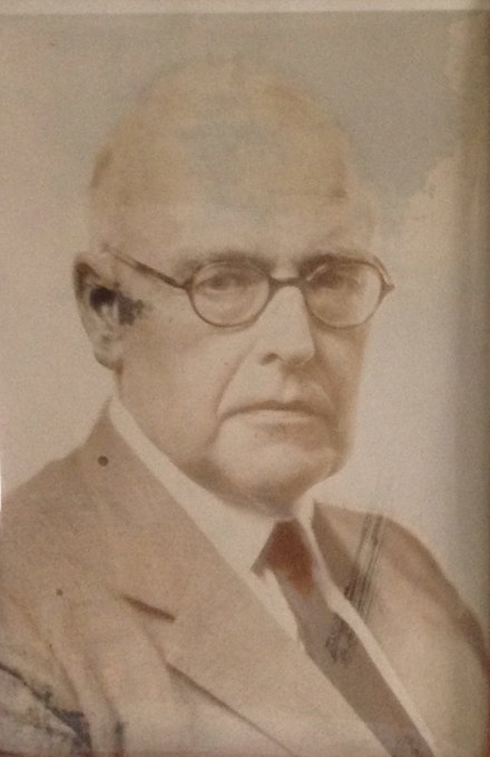 A faded portrait of an older man with glasses in a suit and tie.