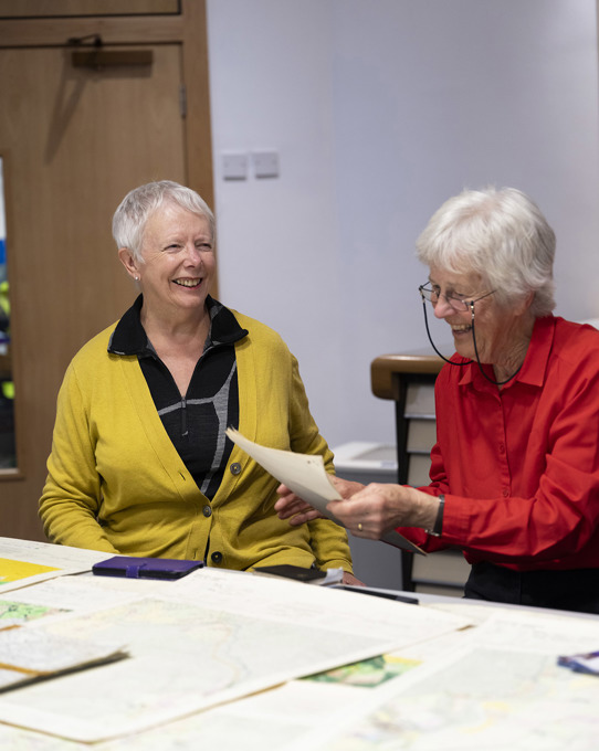 Two women sitting at a table with maps on it.