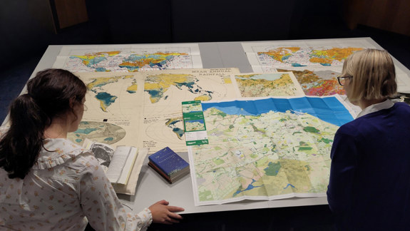 Two people looking at a selection of maps spread out on a table.