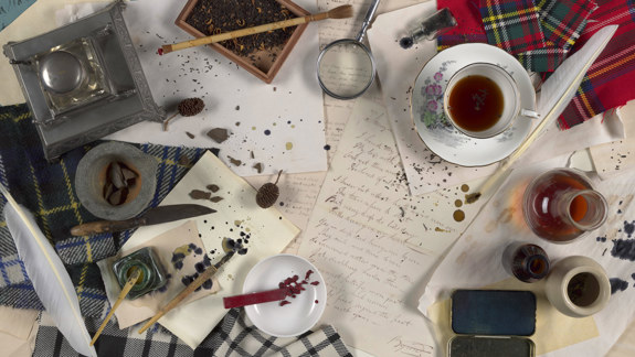 A bird's-eye view manuscripts covered in objects used for creating them, including wax, inkpots, brushes, tartan, and a magnifying glass.