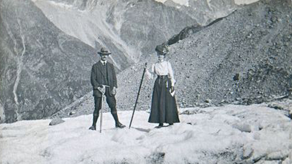 Black-and-white photo of two people standing on a snowy mountain slope, each holding a walking stick, with rugged hills rising in the background.