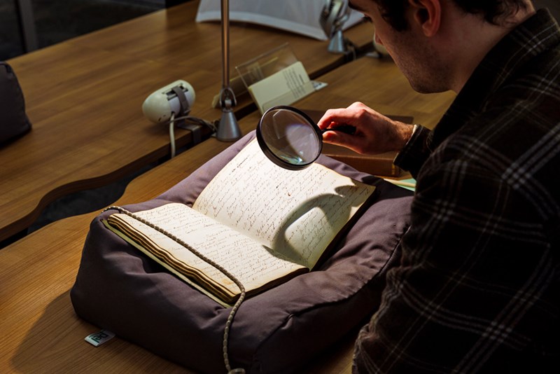 A person sitting at a desk in the Library using a magnifying glass to read a book.
