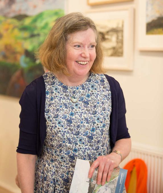Person in a blue and white floral dress holding a book, standing in front of a wall displaying colorful framed artworks.