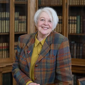 A photograph of Liz Lochhead standing next to a desk in a room with books.