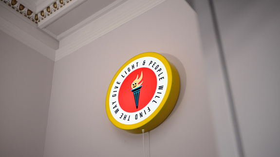A flame framed by a circle with the words 'Give light and people will find the way'.