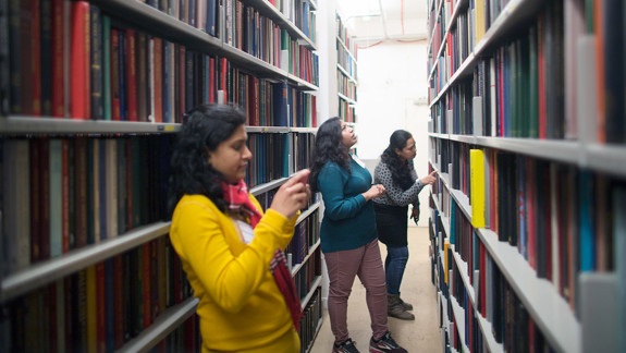 Three adults standing in the stacks looking at books on bookshelves.