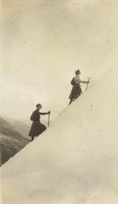 Two women in long skirts climbing up a steep snowy mountain
