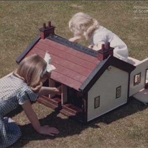 Two young girls playing with a doll's house in a garden on a sunny day.