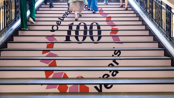 Several people walking up the main Library stairs which have the Library's centenary logo on it.