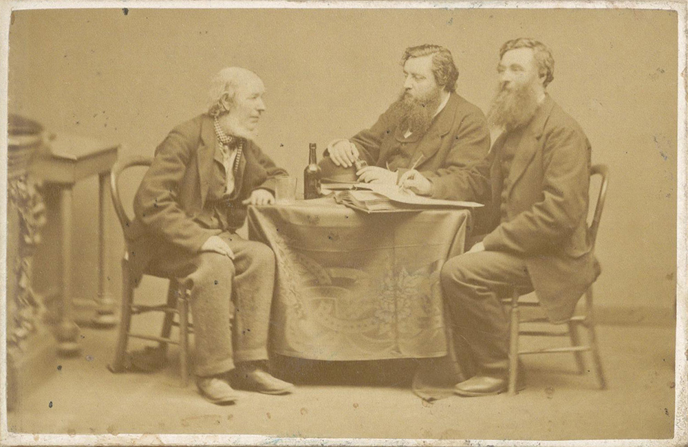 An old photo of three men sitting at a table, one taking notes.