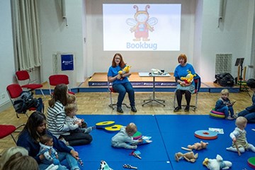 Two people sitting on chairs holding bookbug soft toys. Other people with babies are sitting on blue mats on the floor.