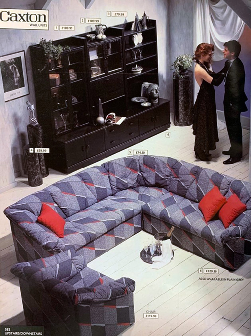 Page from a vintage shopping catalogue showing a large grey and red corner sofa in front of black wall furniture and a man and a woman stand in the corner smiling.