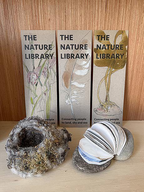 Three bookmarks with botanical drawings stand behind a small next and two pebbles with pages folding out between them