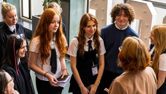 A group of school students stand around listening to an adult leading a group.