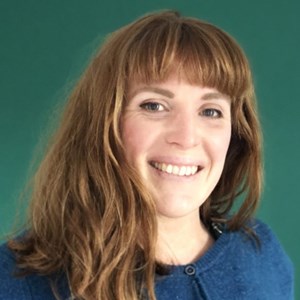 A photo of a woman with brown hair and wearing a blue cardigan. She is looking into the camera and smiling. There is a green background.