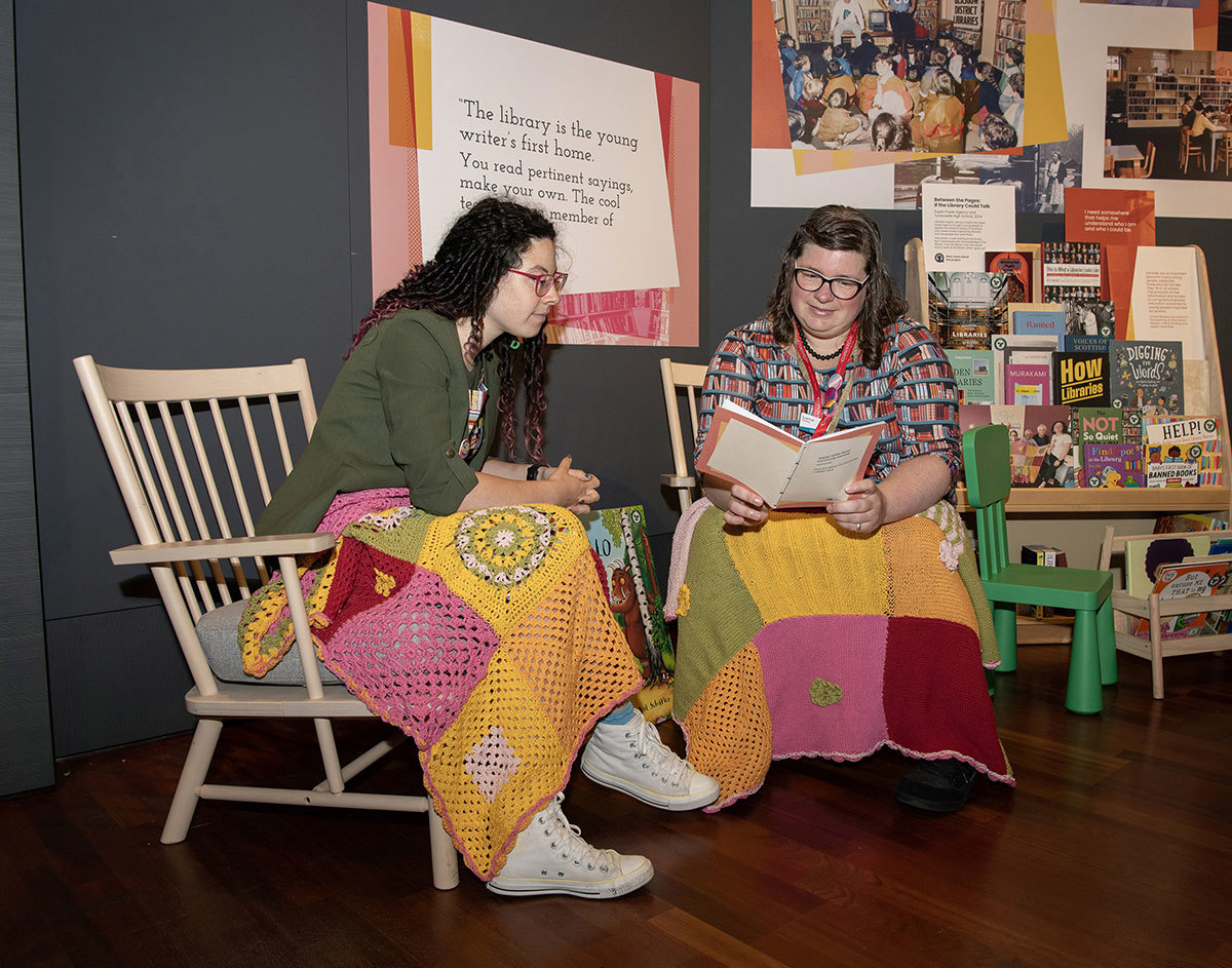 Two people sitting on chairs with rugs over their laps reading a book.