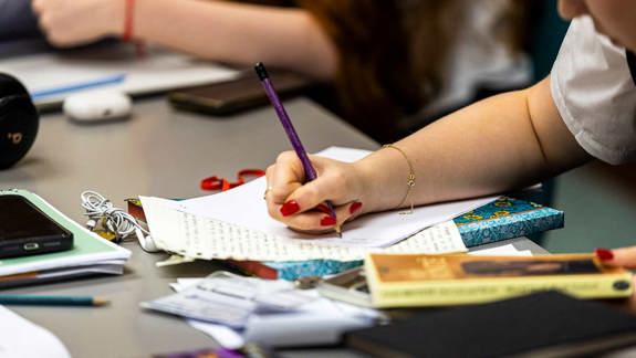 A student leaning over a piece of paper, about to start writing. The desk they are working on is covered in books, notepads, and ear buds.